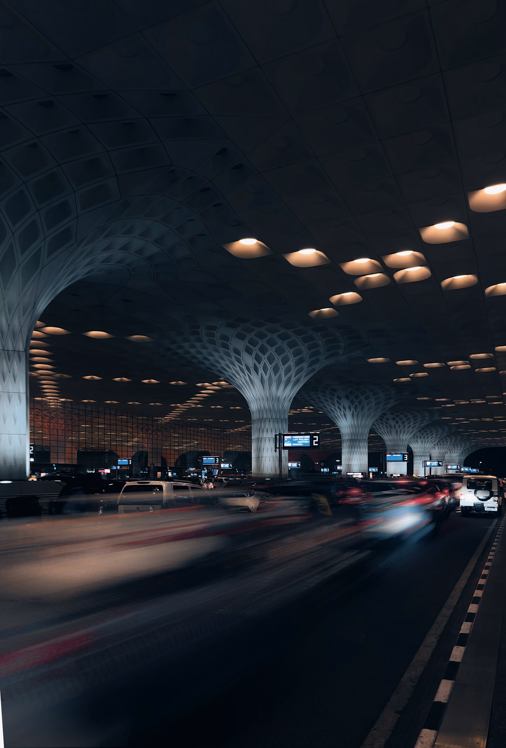 Airport terminal departure roadway at night under a canopy with mushroom-shaped columns, recessed lights, and motion-blurred cars