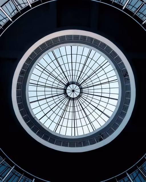 View looking straight up through a circular glass skylight dome with radiating black window bars, framed by concentric round balconies of a spiral atrium.