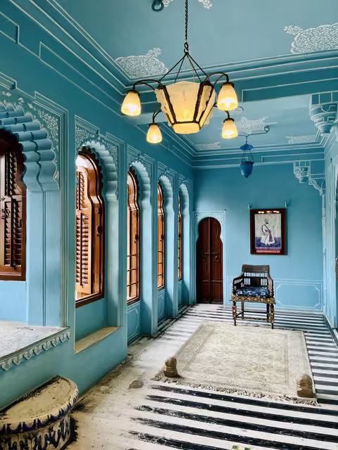 Turquoise painted palace room with cusped arch windows, wooden shutters, glass chandelier, carved chair, framed portrait, striped floor and rug.