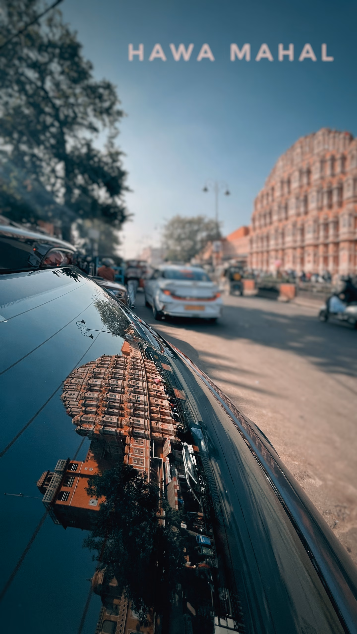 Pink sandstone palace facade reflected in a dark car hood beside a street with cars, scooters and trees, text "HAWA MAHAL" overhead.