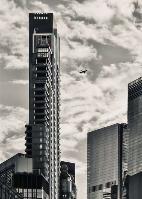 Black and white view of a slender high-rise tower with stacked balconies, glass office blocks, and a small airplane in cloudy sky.