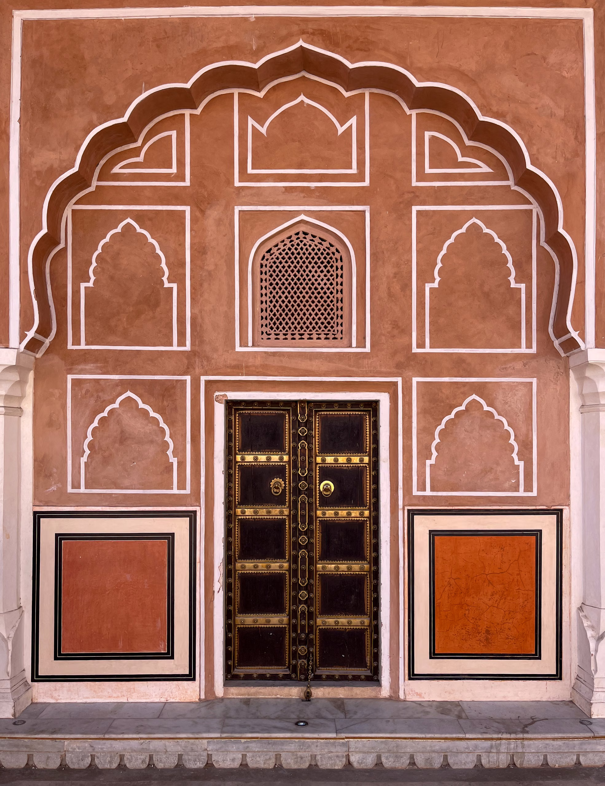 Terracotta wall with white-outlined scalloped arch, brass-studded dark wooden double doors, lattice window above, and two framed painted panels