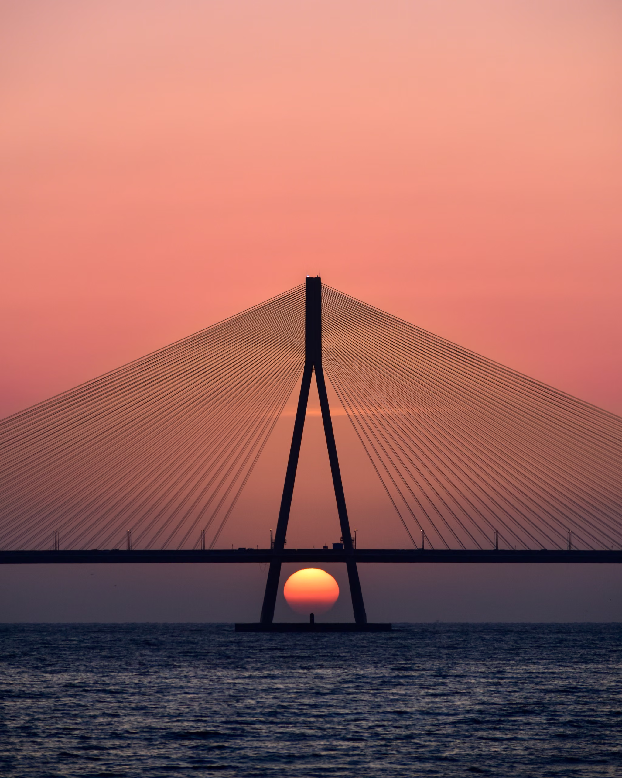 Silhouetted cable-stayed bridge pylon over water with the setting sun framed between its two legs beneath the deck.