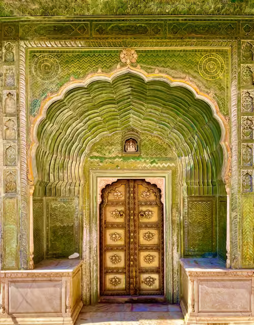 Ornate green carved gateway with scalloped arch, brass-studded wooden double doors, marble benches and small deity niche above.