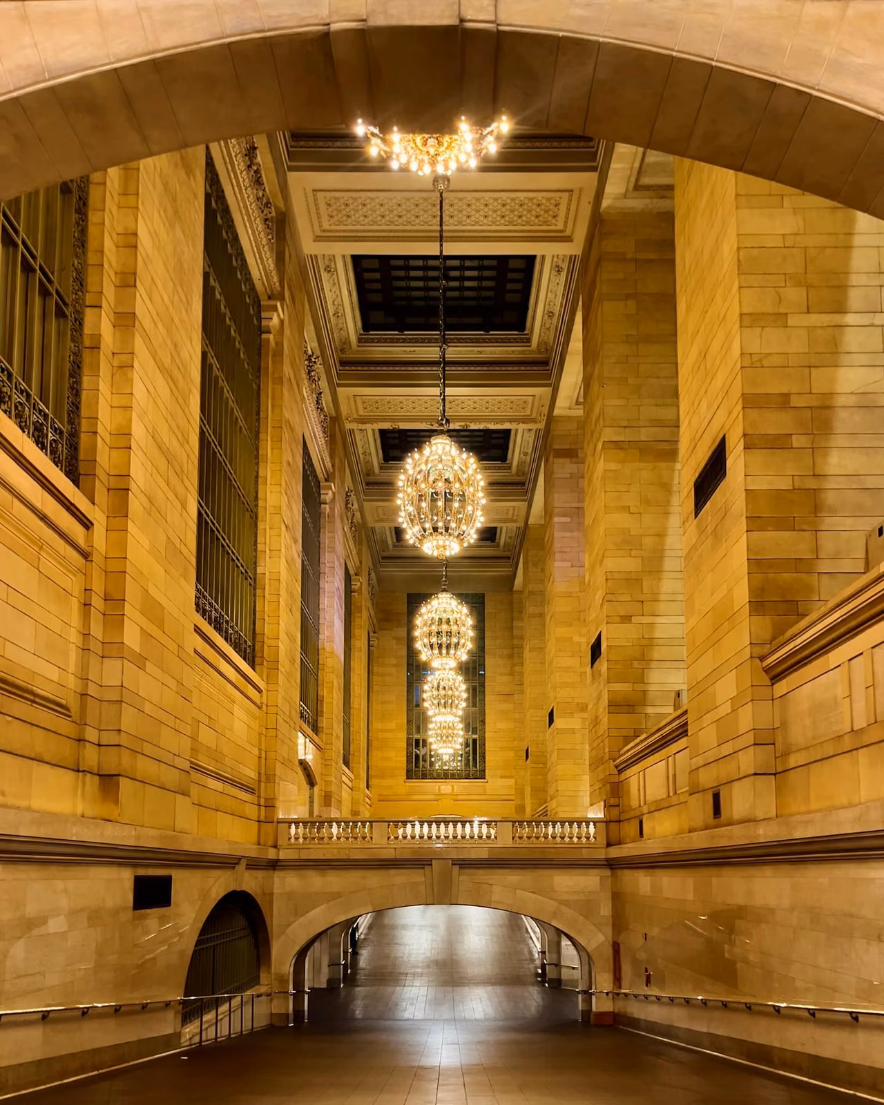 Ornate stone corridor with three tiered gold chandeliers hanging from arched ceiling, leading to a lower tunnel with a stone balustrade.