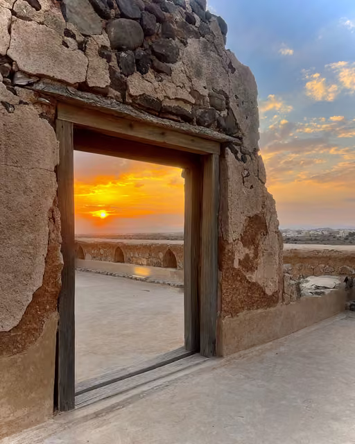 Weathered stone and mud-brick wall with a wooden-framed doorway opening onto a terrace, sun setting over desert plain beyond