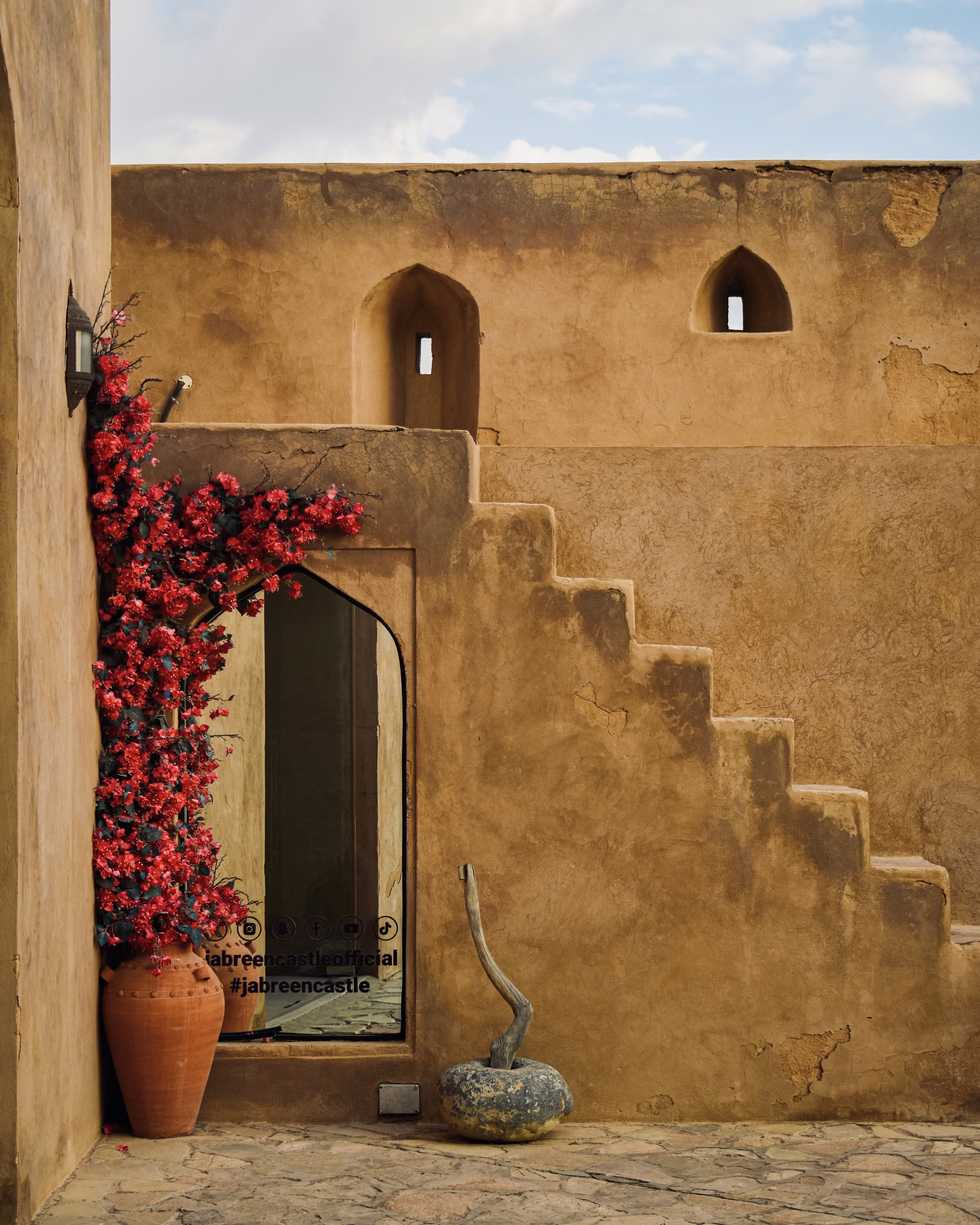 Ochre mud-plaster courtyard wall with pointed arch doorway, red bougainvillea in a clay urn, stepped stairs, and slit windows.