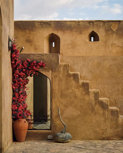 Ochre mud-plaster courtyard wall with pointed arch doorway, red bougainvillea in a clay urn, stepped stairs, and slit windows.
