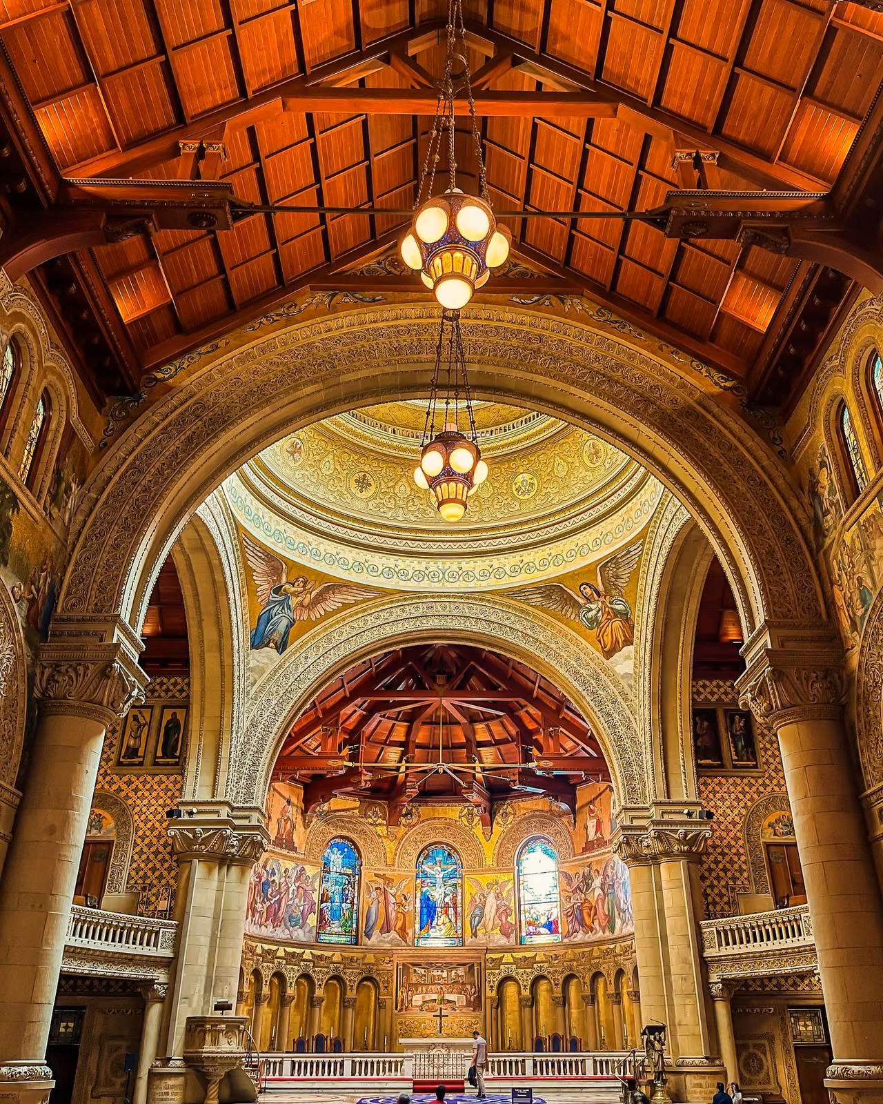 Ornate church interior with wooden vaulted ceiling, gilded dome, arched apse with stained glass windows and mosaic angel murals, hanging chandelier lamps.