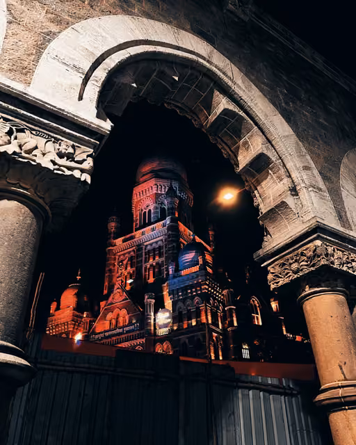 Floodlit Gothic municipal building with domed tower seen at night through a carved stone arch, orange and blue lighting, corrugated hoarding below