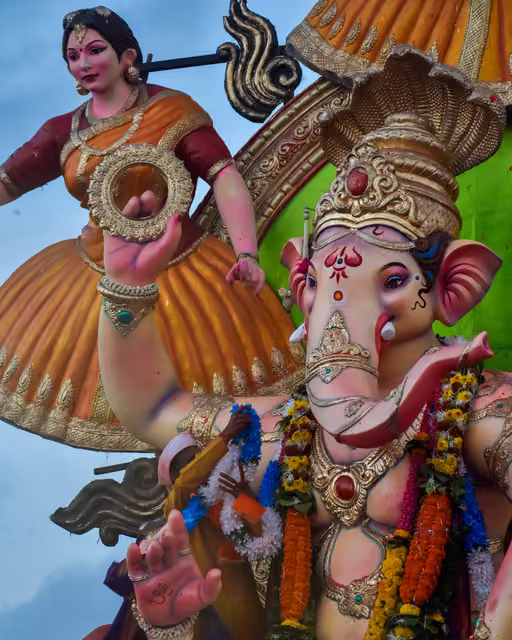 Large painted Ganesha idol with gold crown and flower garlands beside a female figure in orange sari, against pale sky