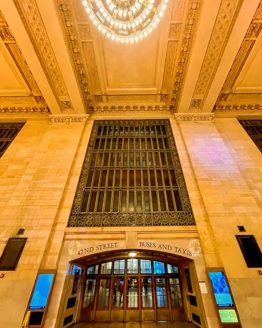 Ornate marble entrance hall with tall arched window grille, carved ceiling, chandelier, and doors labeled '42nd Street Buses and Taxis'.