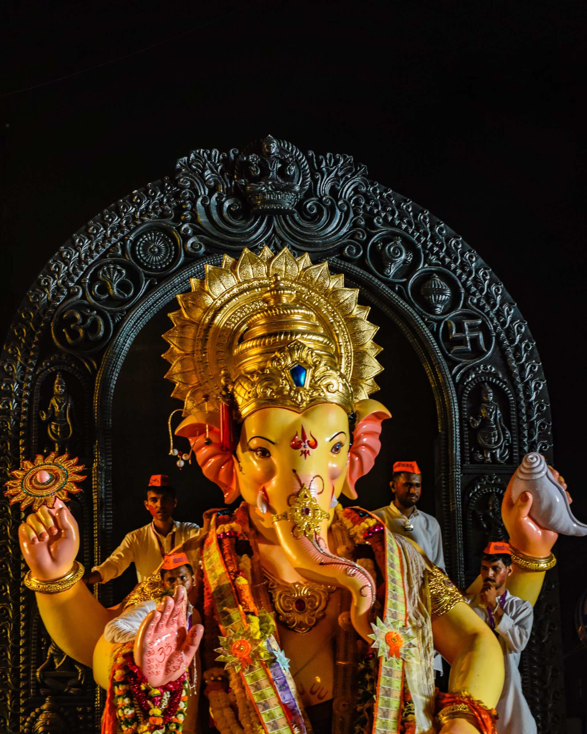 Large four-armed Ganesha idol with gold crown, garlands and raised palms, framed by carved dark arch; men in white shirts and orange caps stand behind.