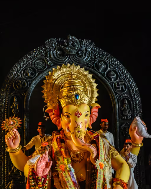 Large four-armed Ganesha idol with gold crown, garlands and raised palms, framed by carved dark arch; men in white shirts and orange caps stand behind.