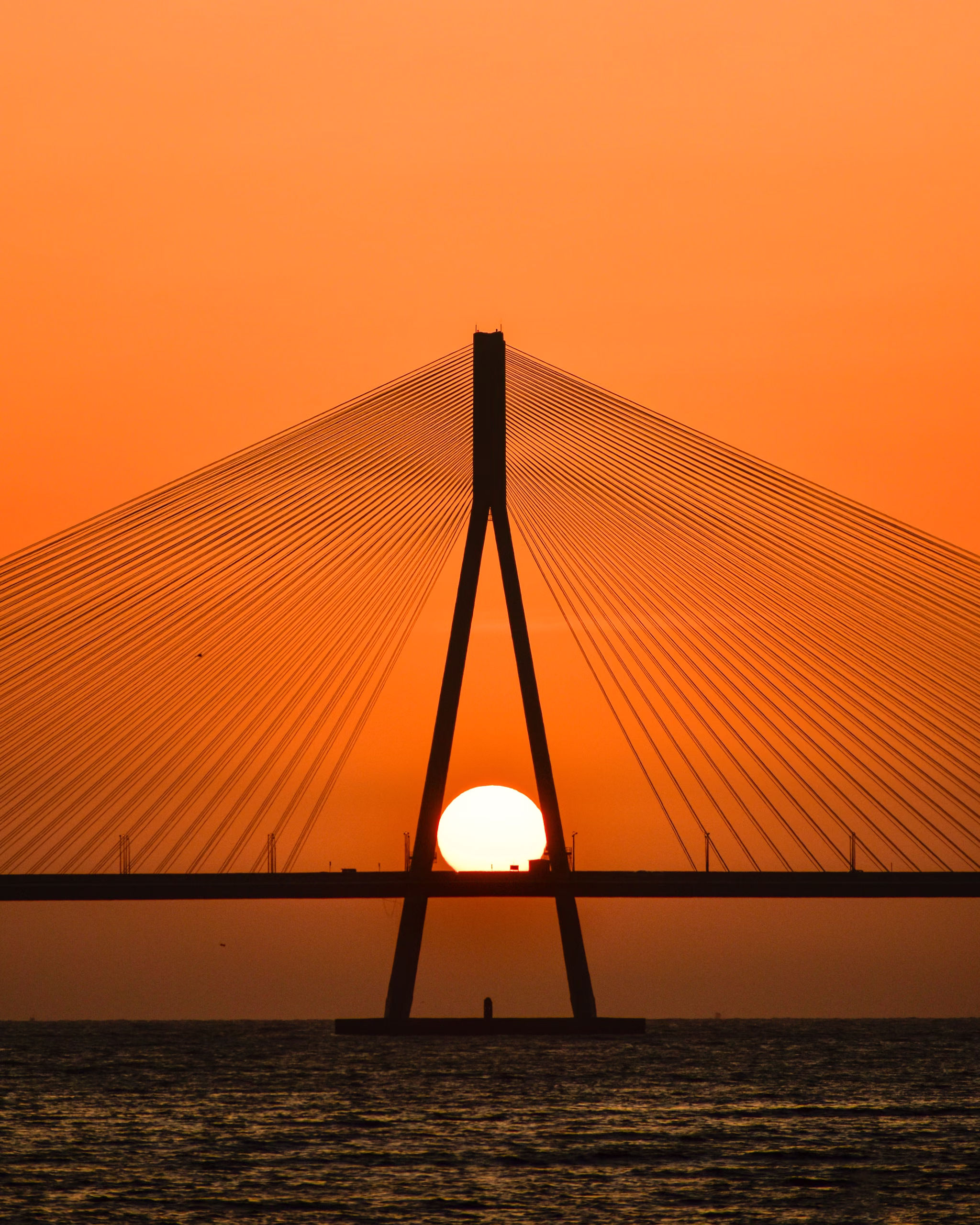 Silhouetted cable-stayed bridge pylon over water, the sun setting directly between its two angled legs against an orange sky