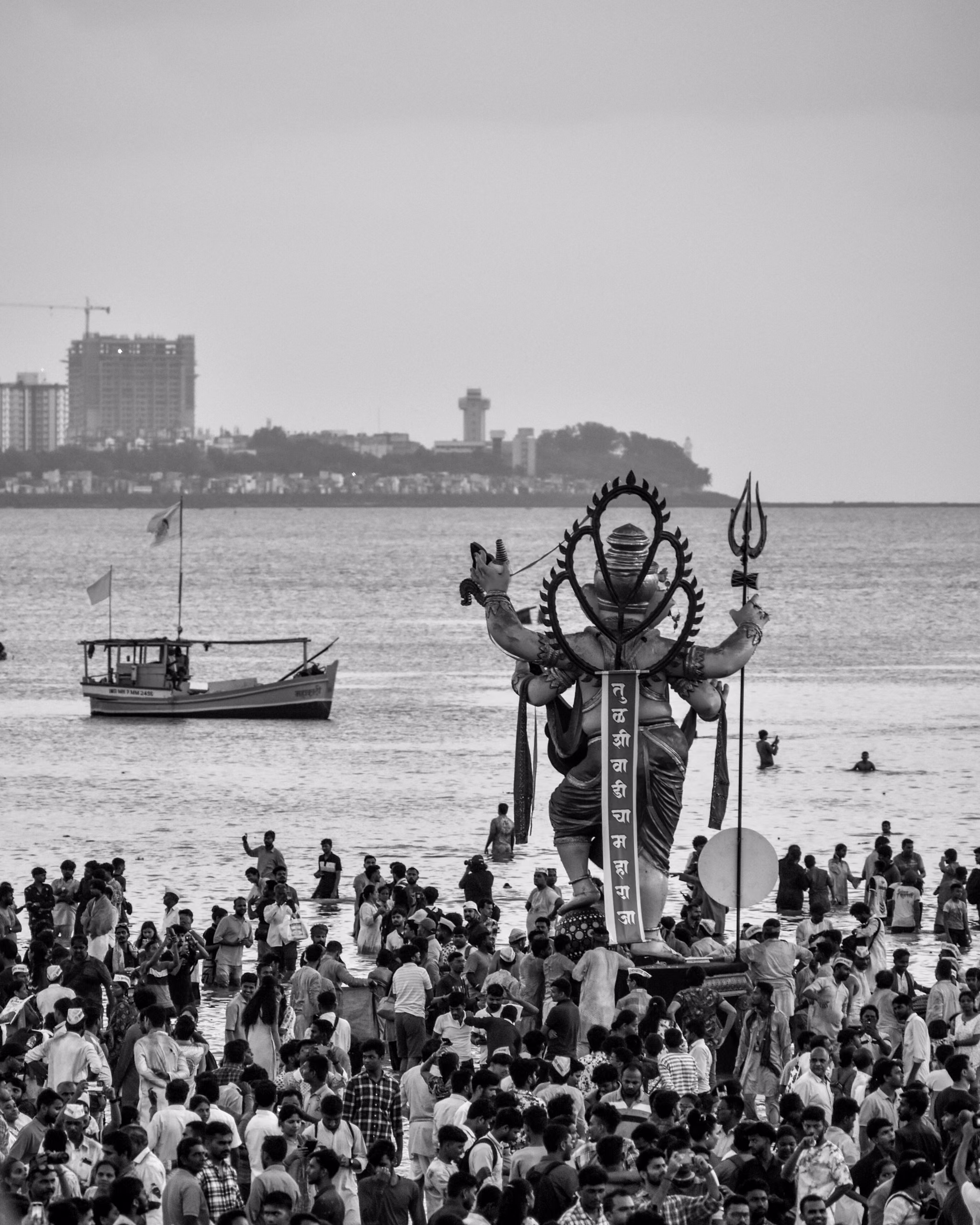 Black-and-white view of a large Ganesh idol carried into the sea, surrounded by a dense crowd, with a boat and city skyline behind.