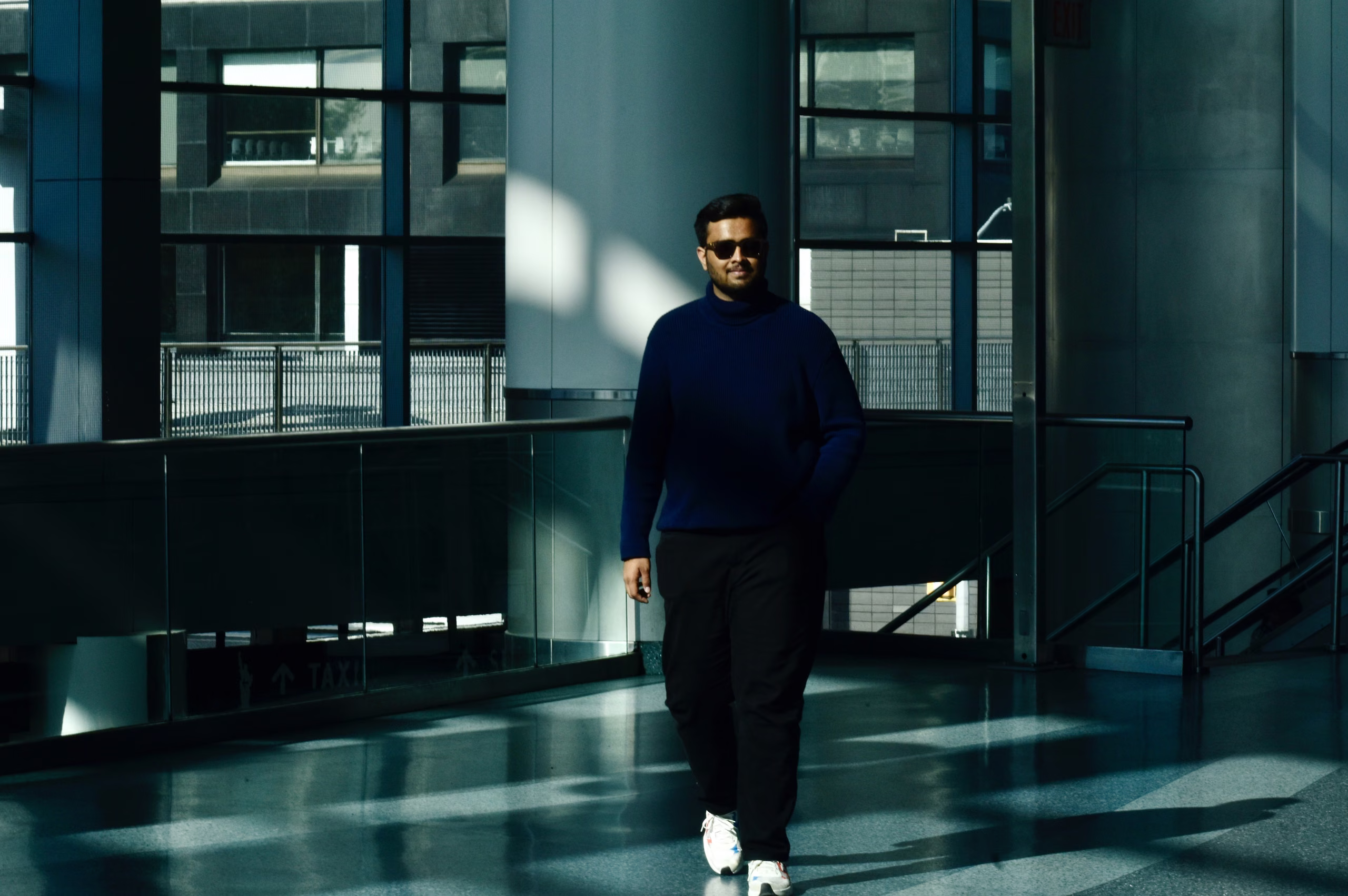 Man in navy turtleneck sweater, dark trousers, white sneakers and sunglasses walks through a glass-walled terminal walkway in sunlight.