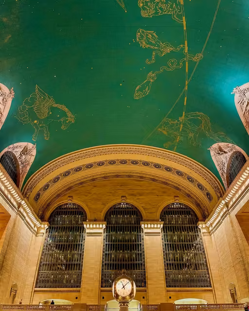 View of Grand Central Terminal's arched windows and gold clock, beneath a green ceiling painted with gold zodiac constellations.