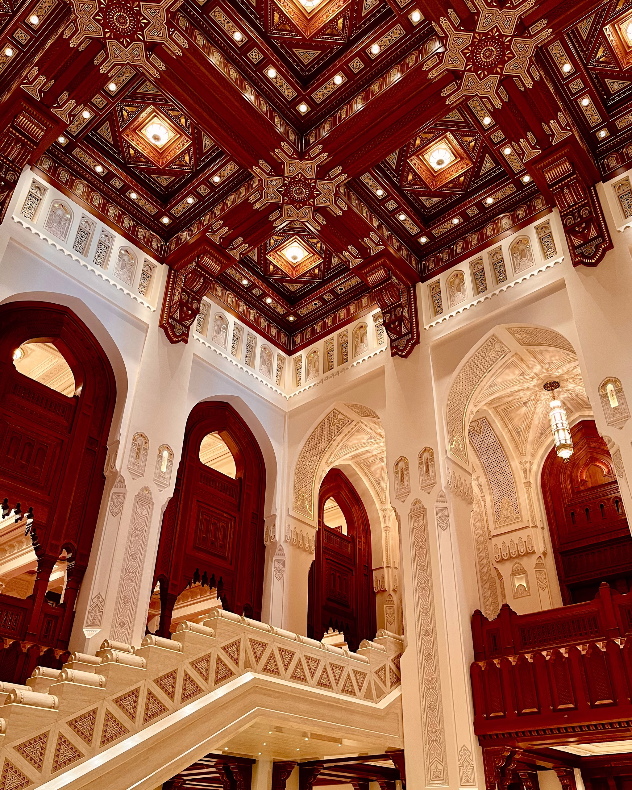 Ornate interior atrium with coffered dark red wooden ceiling, recessed lights, white carved plaster walls, pointed arches and pale stone staircase.