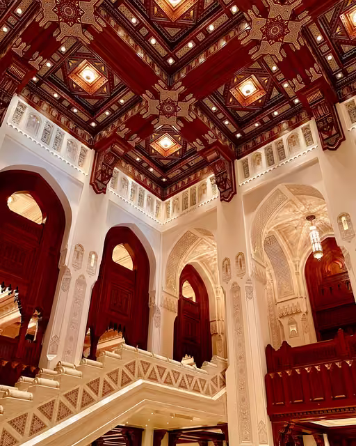 Ornate interior atrium with coffered dark red wooden ceiling, recessed lights, white carved plaster walls, pointed arches and pale stone staircase.