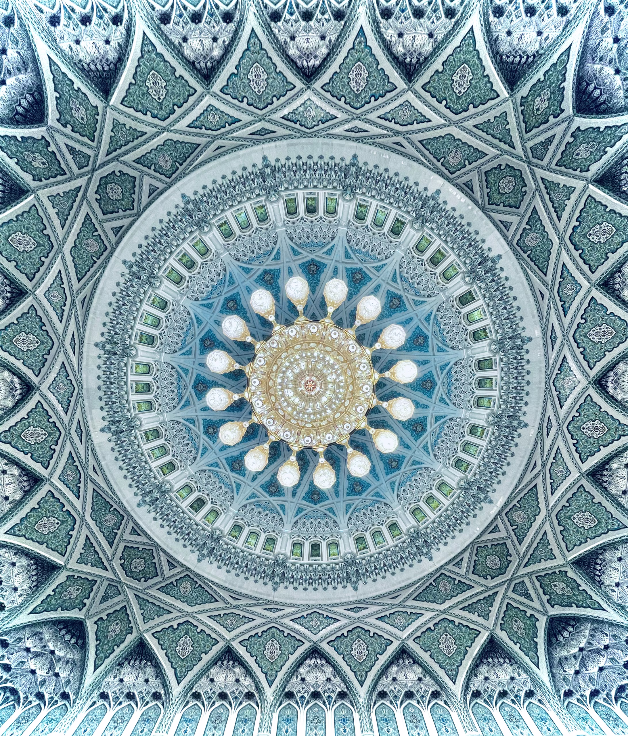 Upward view of a mosque dome: teal and white geometric tilework, arched clerestory windows, and a large gold crystal chandelier with sixteen radiating lamps.